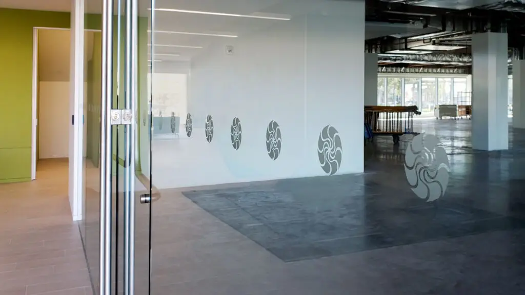 A modern office space with glass partitions featuring circular frosted decals, leading to an open area with exposed ceilings and large windows.