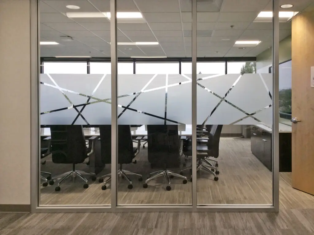 Glass-walled conference room with frosted geometric decals, black office chairs, a long table, and large windows in the background.