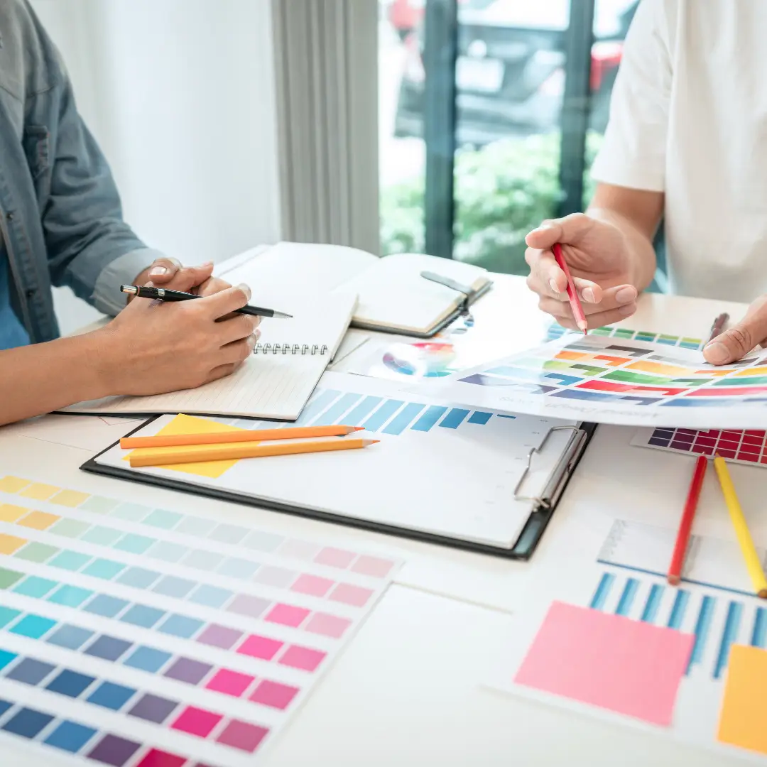 Two people reviewing color swatches and design samples, collaborating on a creative project with notebooks and pens on the table