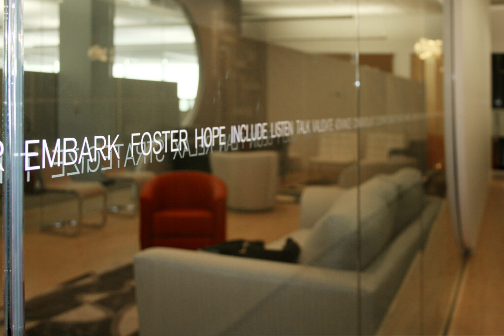 A glass office partition with motivational words reflects a modern lounge with sofas, chairs, and warm lighting.