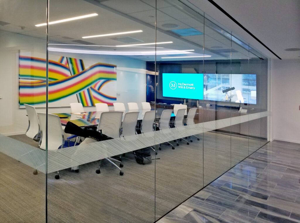 Modern glass-walled conference room with a frosted diagonal stripe decal, a long table surrounded by white office chairs, a large screen displaying "McDermott Will & Emery," and a vibrant abstract mural in the background.